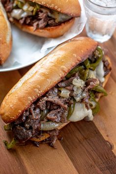 two sandwiches with meat, peppers and onions on a wooden table next to a glass of water