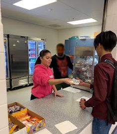 two people standing at a counter in a store with food on it and one person handing something to another person