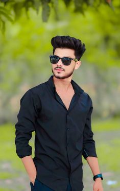 a man with sunglasses on standing in front of a green tree and wearing a black shirt