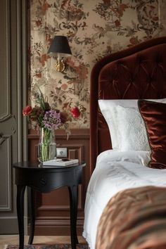 a small table with flowers on it in front of a bed and wallpapered walls