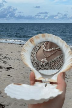 a person holding up a shell with jewelry in it on the beach next to the ocean