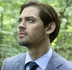 a man in a suit and tie looking off into the distance with trees behind him