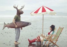 a woman sitting in a chair next to a giant fish on the beach with an umbrella