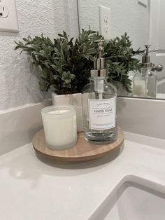 a candle sits on a wooden tray in front of a bathroom sink with a plant and soap dispenser