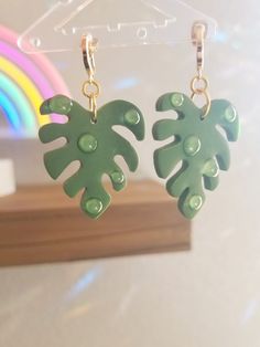 two green leaf shaped earrings hanging from a hook on a white wall with rainbow in the background