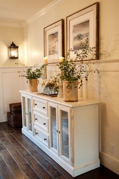 two framed pictures on the wall next to a white cabinet with drawers and vases filled with flowers