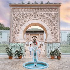 two people are walking in front of an archway with a fountain and potted plants