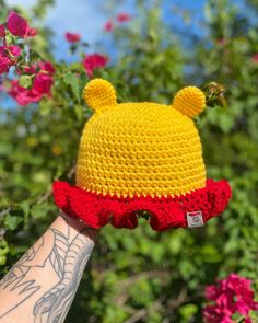 a hand holding up a crocheted winnie the pooh hat in front of some flowers