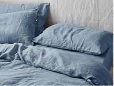 an unmade bed with blue linens and pillows on the headboard, in front of a white wall