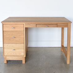 a wooden desk with three drawers in front of a white wall and concrete flooring