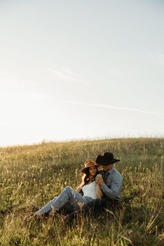 a man and woman sitting in the grass with their arms around each other, wearing cowboy hats