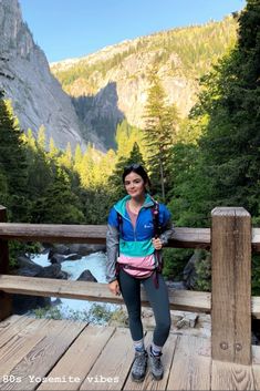 a woman standing on a wooden bridge next to a river and mountains in the background