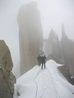 two people walking up a snow covered mountain in the fog with rocks on either side