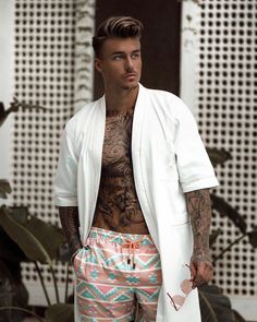 a man with tattoos wearing colorful shorts and a white jacket standing in front of plants