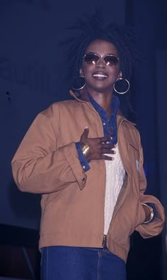 a woman with dreadlocks standing in front of a blue wall wearing a brown jacket