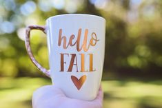 a hand holding a coffee mug with the words hello fall painted on it in gold