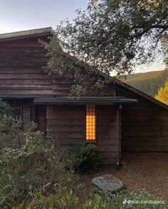 an old log cabin with a window lit up in the evening light, surrounded by trees and shrubs