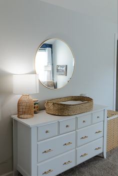 a white dresser with a basket on top and a round mirror above it in a bedroom