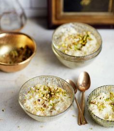 three glass bowls filled with rice and nuts next to two spoons on a table