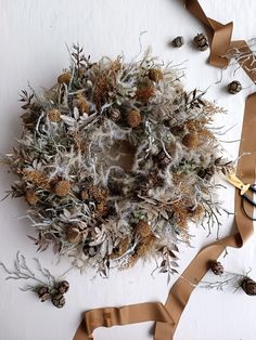 a wreath made out of dried plants and pine cones on a white surface with ribbon