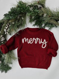 a red sweater with merry written on it next to pine branches and white berries in the background