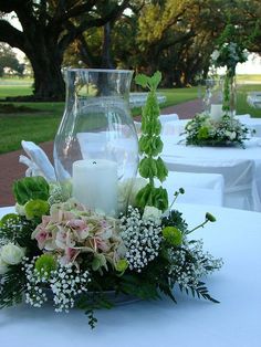 a vase with flowers and candles on a white table cloth at an outdoor wedding reception