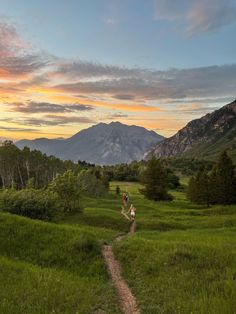 two people walking down a trail in the middle of a grassy field with mountains in the background