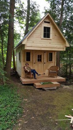 a small cabin in the woods with a man sitting on a chair next to it