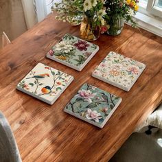 four square coasters with flowers on them sitting on a wooden table next to a vase