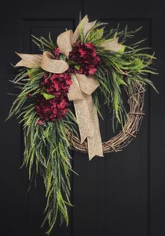 a wreath with red flowers and greenery hanging on a black front door decorated with burlap ribbon