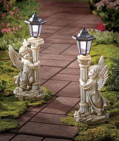 two angel statues sitting next to each other on top of a brick walkway in front of flowers