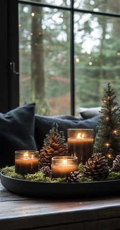 candles are lit on a tray with pine cones and evergreens in front of a window