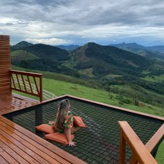 a woman sitting on top of a wooden deck next to a lush green hillside under a cloudy sky