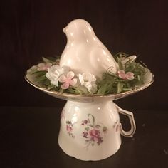 a white bird figurine sitting on top of a bowl filled with flowers and greenery