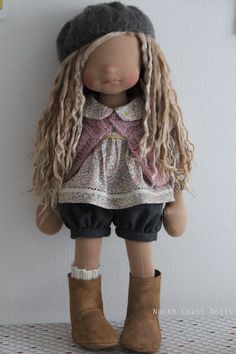 a doll with long blonde hair wearing boots and a hat is standing against a wall