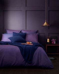 a bed with purple sheets and pillows in a dark colored room next to a lamp