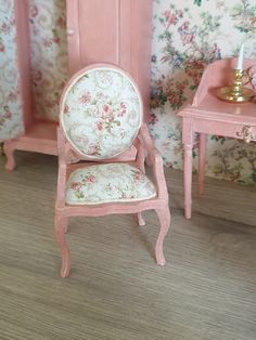 a pink dollhouse with a chair and table next to it in front of a floral wallpapered wall