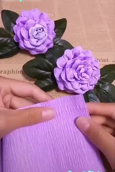 two hands are holding purple paper and some flowers