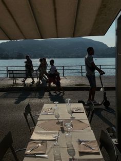 several people are walking by the water near tables with place settings on them and one person is riding a scooter