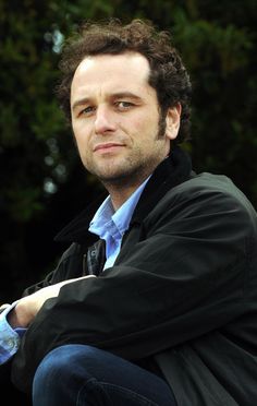 a man with curly hair sitting in front of trees and looking at the camera while wearing a black jacket