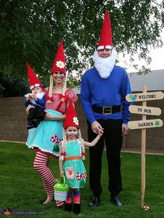 three people dressed up as gnomes standing in the grass next to a wooden sign