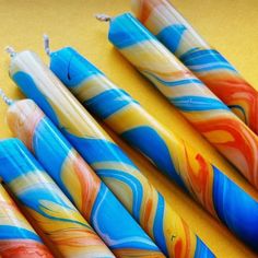 four colorful candles are lined up on a yellow table top, one is blue and the other has orange swirls