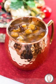 a copper mug filled with soup sitting on top of a red table cloth next to christmas decorations