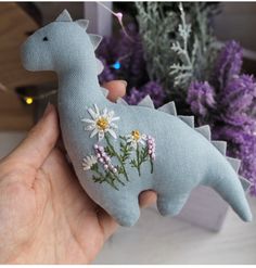 a hand holding a stuffed toy dinosaur with daisies on it's back and flowers in the background