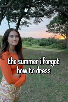 a woman in an orange top and floral skirt standing next to trees with her arms crossed