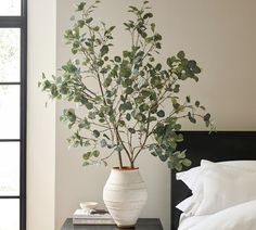 a white vase filled with green leaves on top of a table next to a bed