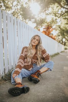 a woman sitting on the ground next to a white fence wearing black boots and jeans