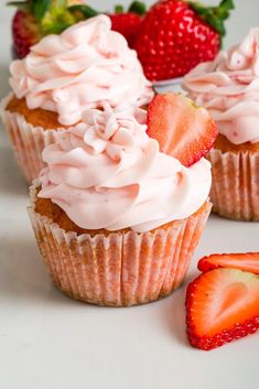 three cupcakes with frosting and strawberries next to them on a white surface