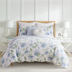 a white bed with blue and green flowers on the comforter, pillows and lamps