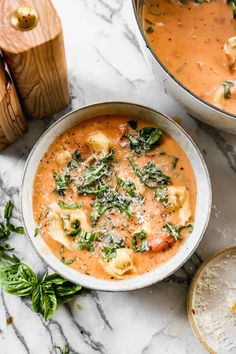 two bowls of pasta and spinach soup on a marble countertop with basil leaves